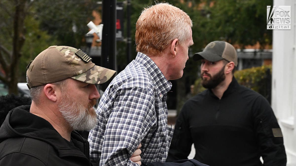 A man wearing a plaid shirt is escorted out of a police vehicle by armed officers.