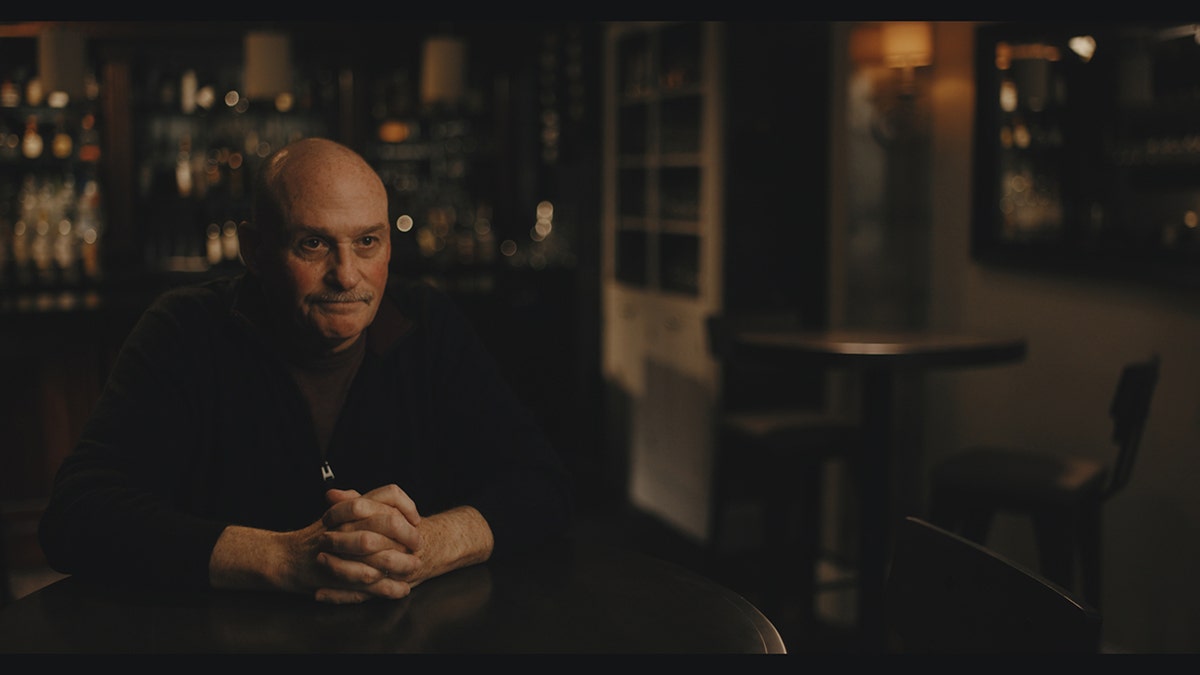 Warren Tobias wearing dark clothing and sitting in front of a wooden table.