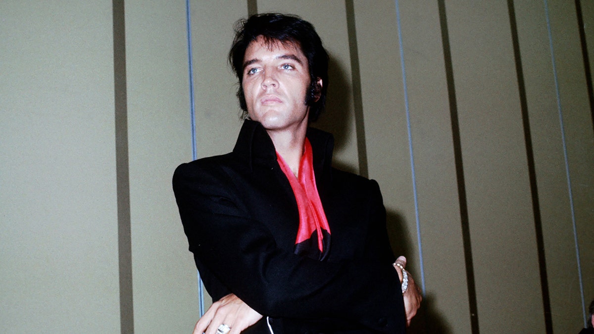 Elvis Presley looks off in the distance wearing a black jumpsuit with a red scarf