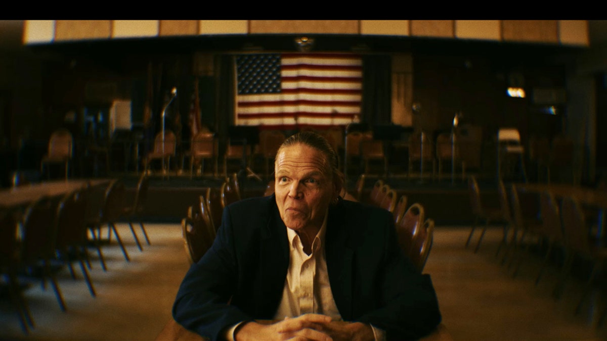 Dan Foote wearing a black blazer and a white shirt sitting with the American flag above his head.