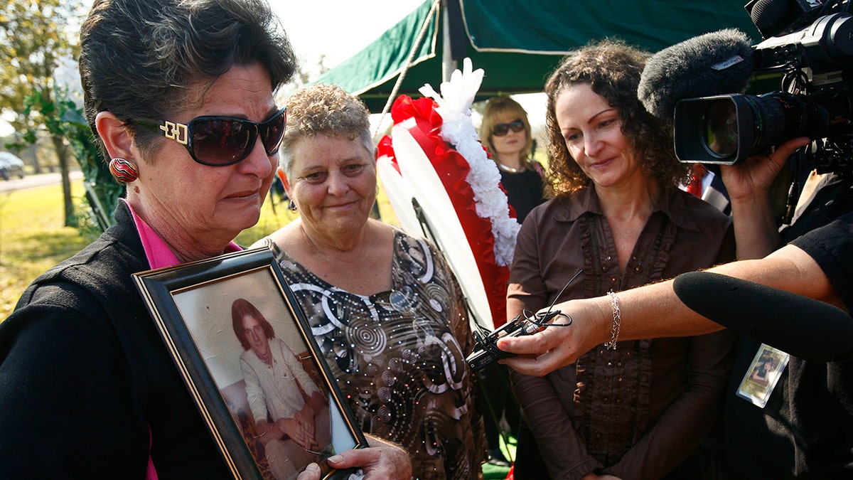 A woman wearing sunglasses looking heartbroken as she holds a photo of a murdered victim.