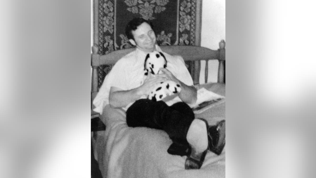 Dean Corll laying on a bed holding a stuffed animal smiling.