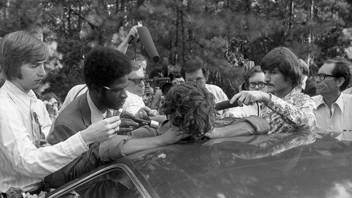 Elmer Wayne Henley covering his face as reporters surround him in front of a car.