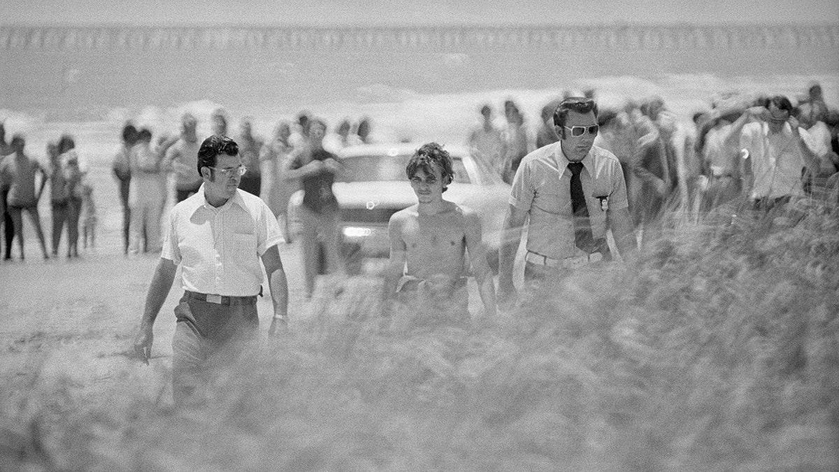 Elmer Wayne Henley walking with police on a beach.
