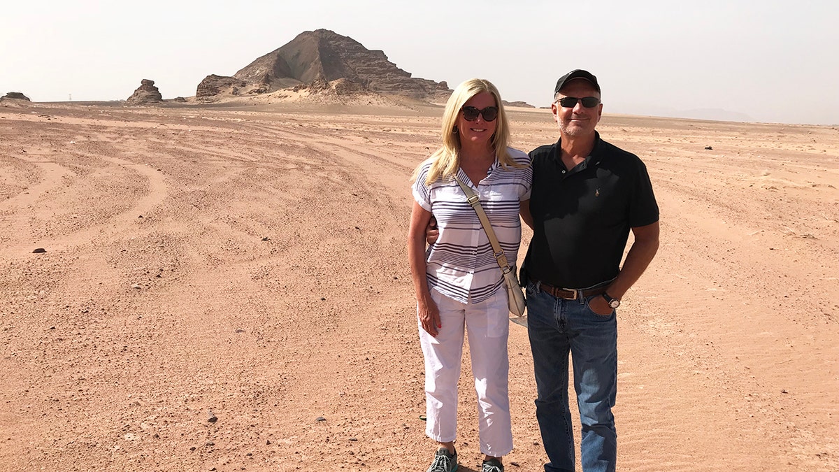 Lori Milliron in a white jumpsuit next to Larry Rudolph in a dark shirt and jeans standing in a desert.