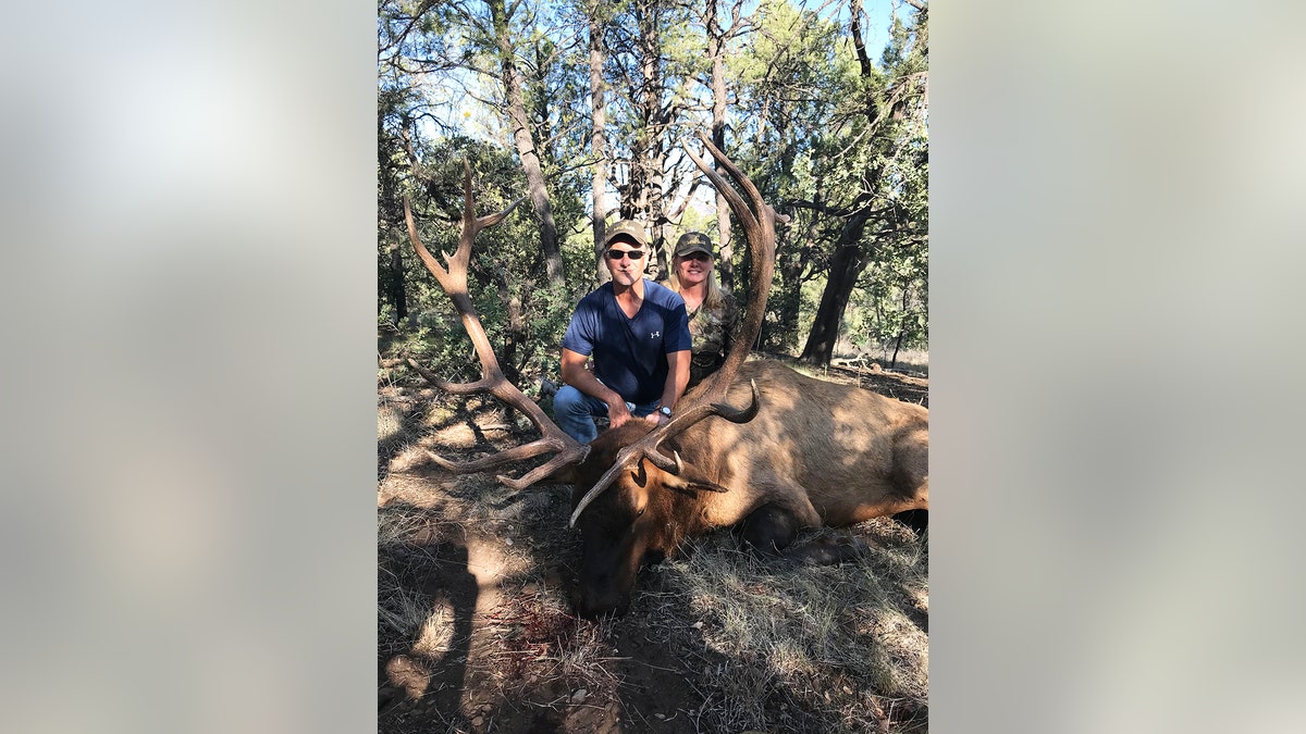 Larry Rudolph wearing a blue shirt and a green hat next to Lori Milliron wearing green as they kneel in front of a elk they hunted.
