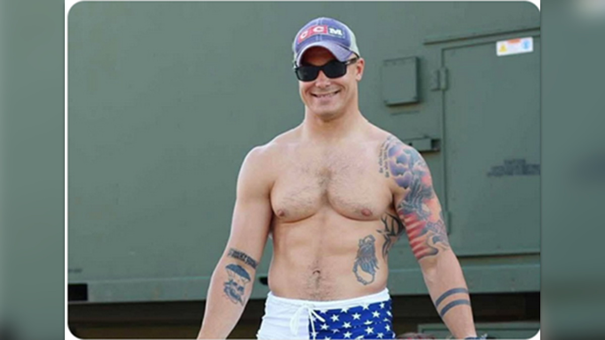 Travis Decker pictured with a clean shave and shirtless, showing tattoos on both arms and his left side