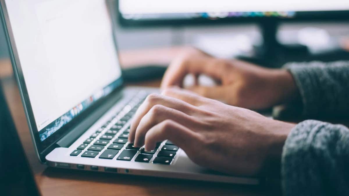 Person typing on a laptop at a desk with computer monitors in the background.