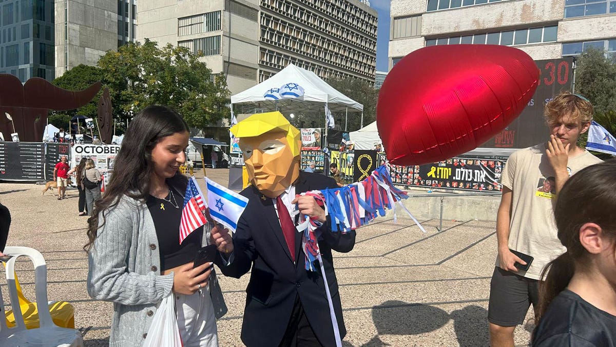Person wearing Trump mask hands U.S. and Israeli flags to a woman in Tel Aviv’s Hostages Square.