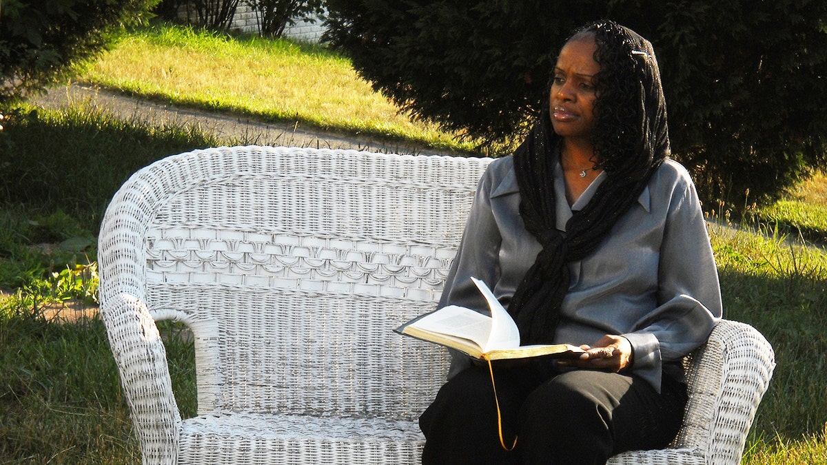 Dr. Mildred Muhammad sitting on a white chair holding a book.