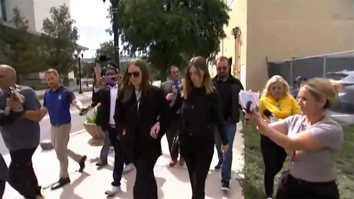 Two people in dark clothing walk together past reporters and photographers outside a courthouse.