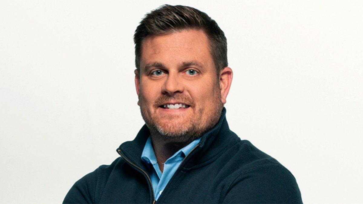 Brian Thompson in a blue button down shirt and blue zip-up smiles for the camera