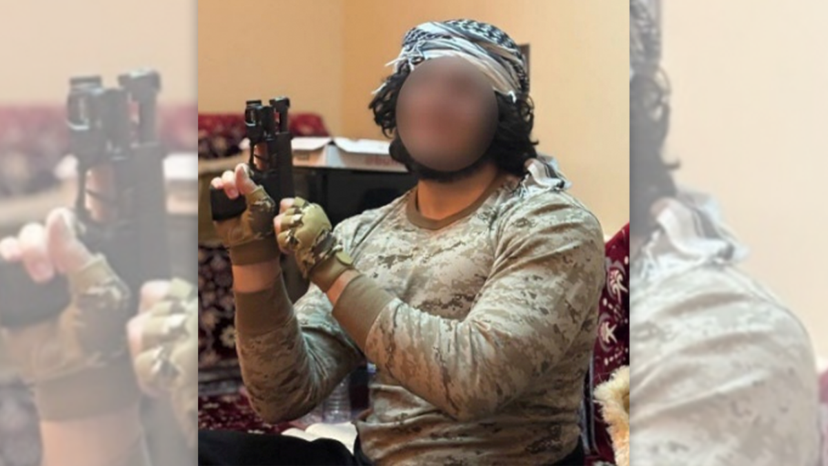 An image of a man whose face has been blurred wearing a keffiyeh on his head and holding a handgun