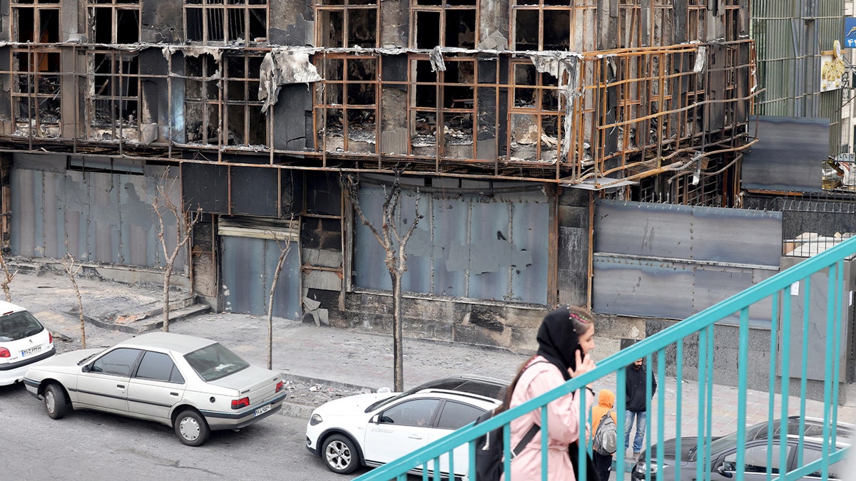 A woman climbs stairs near a heavily damaged structure bearing signs of fire and destruction in an urban area.