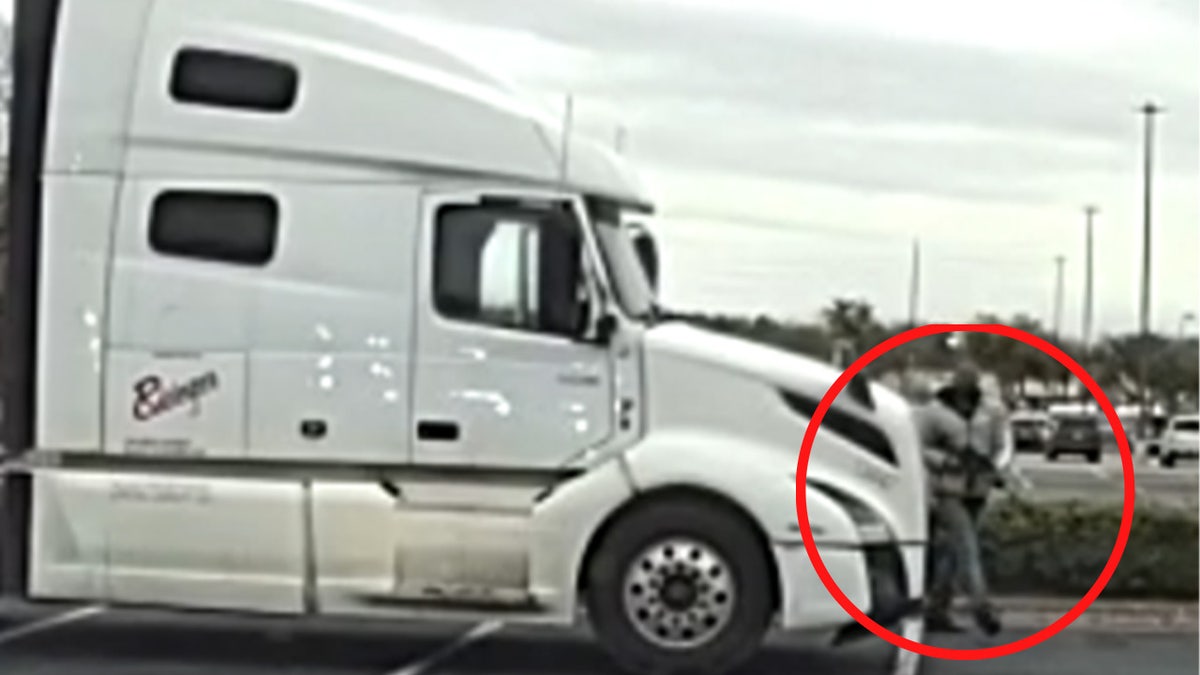 Surveillance image shows a white semi-truck parked in a shopping center lot as a man stands near the front of the vehicle hold a gun.