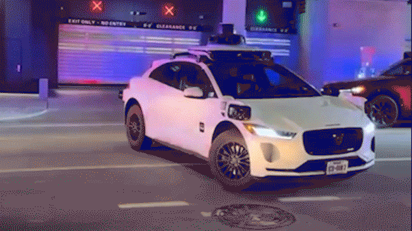 A Waymo autonomous vehicle is parked near police cars with flashing red and blue lights outside a downtown Austin parking facility at night.